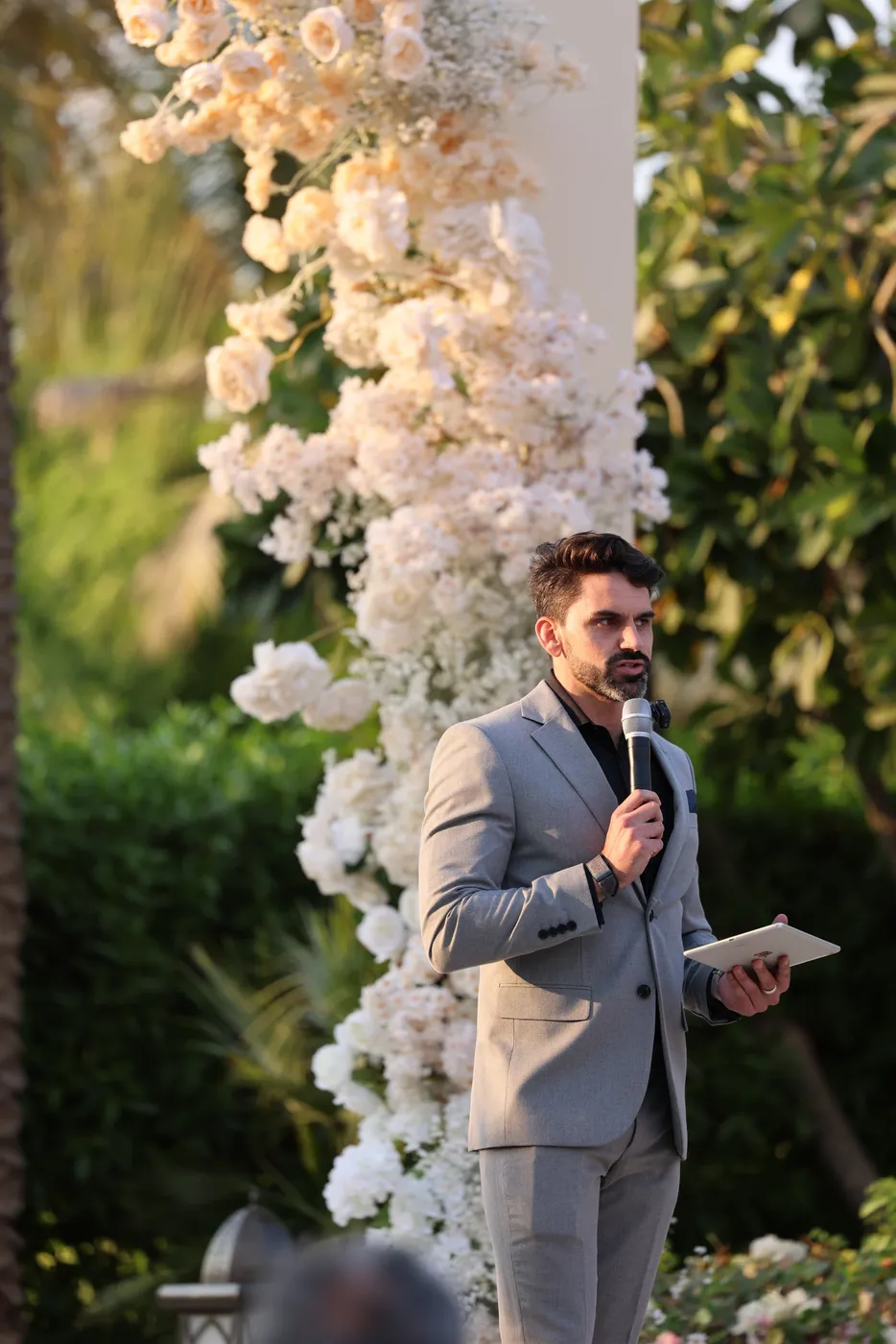 Darryl Rees speaking as wedding celebrant beside a white rose floral arch